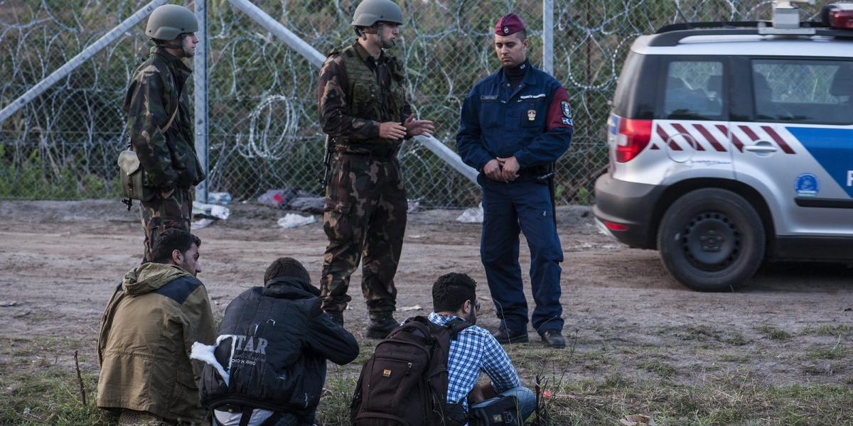 Katonák és rendőrök három elfogott illegális bevándorlót őriznek a biztonsági határzár mellett a szerb-magyar határon, Röszke té