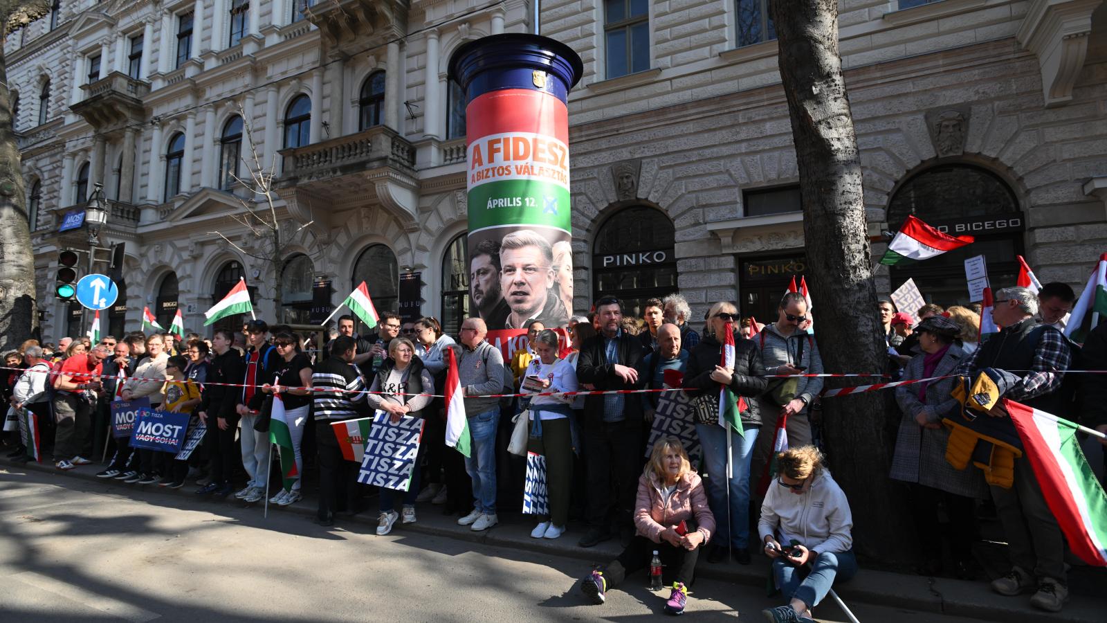 Illusztráció: Tisza-szimpatizánsok várakoznak egy Fidesz-plakát mellett március 15-én